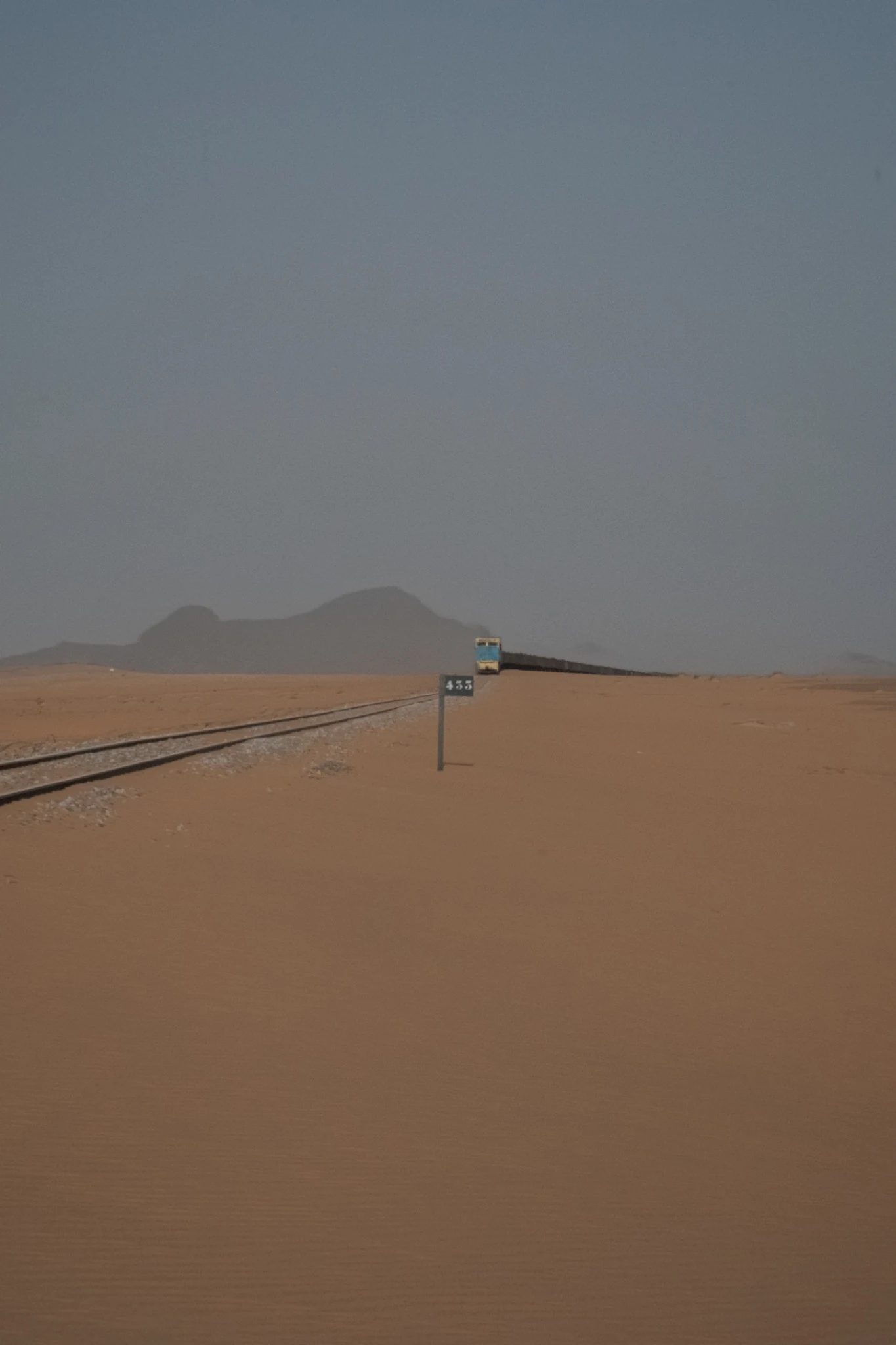 Railway tracks running through sand towards distant rock formations and a train.