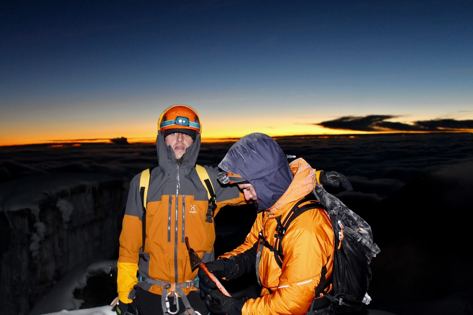 Two climbers wearing helmets and harnesses, with the first light above the cloud behind them.