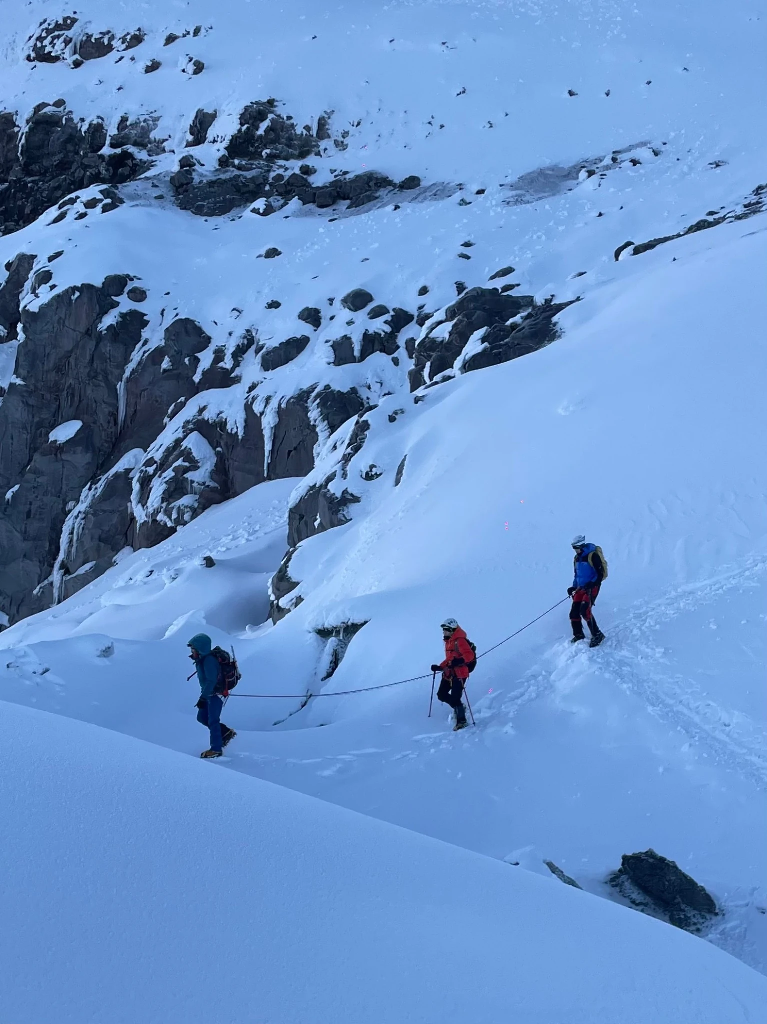 Three climbers linked by a rope on a snow slope beneath exposed rock.