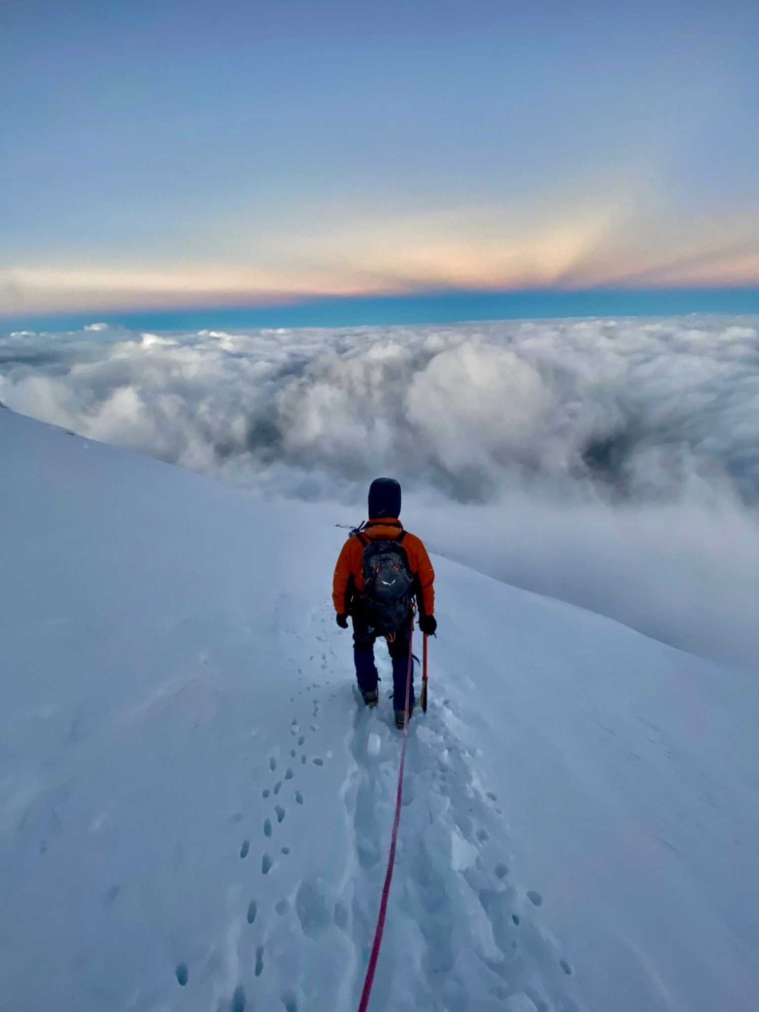 A climber following footprints above a bank of cloud, with a rope running towards the camera.