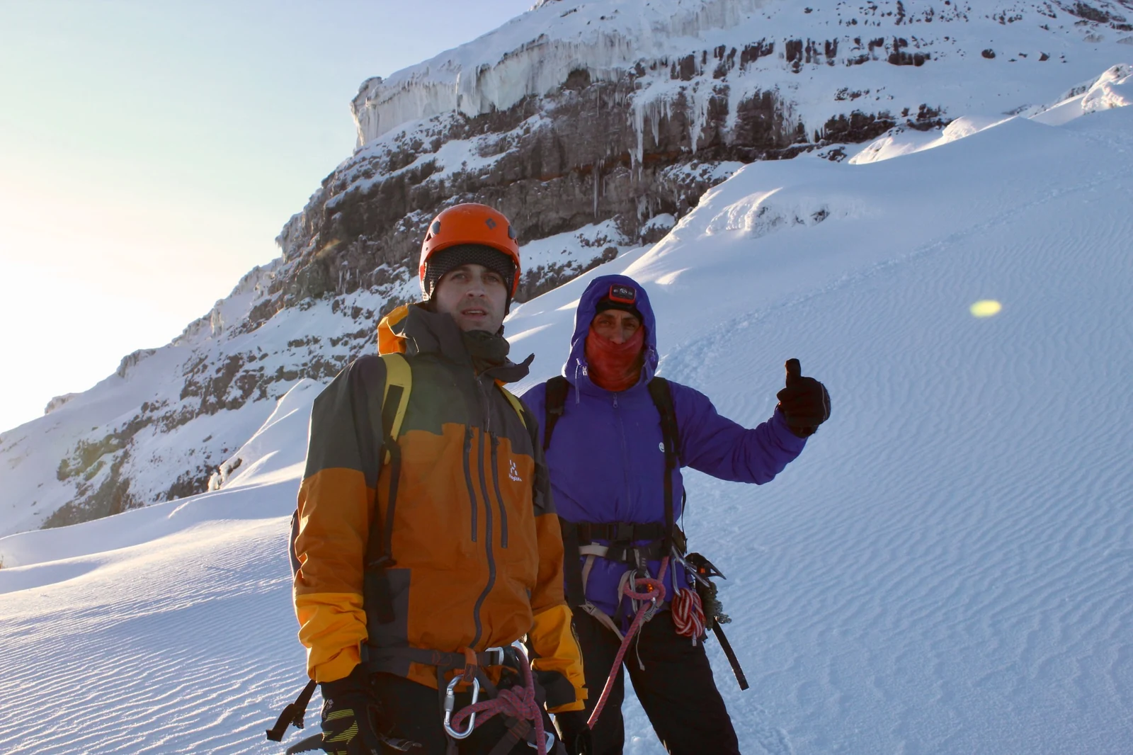Two climbers standing on snow beneath a rock face edged with ice.