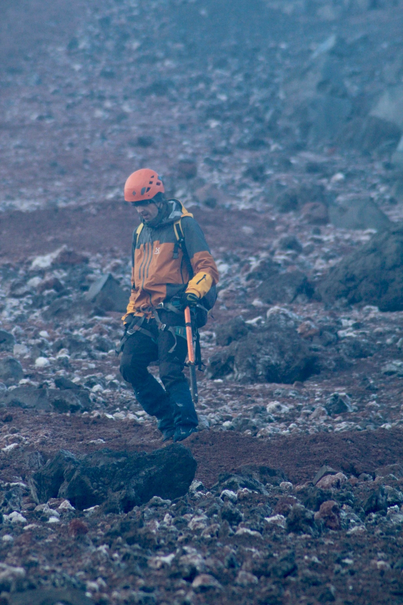 A climber wearing an orange helmet standing among loose volcanic rocks.