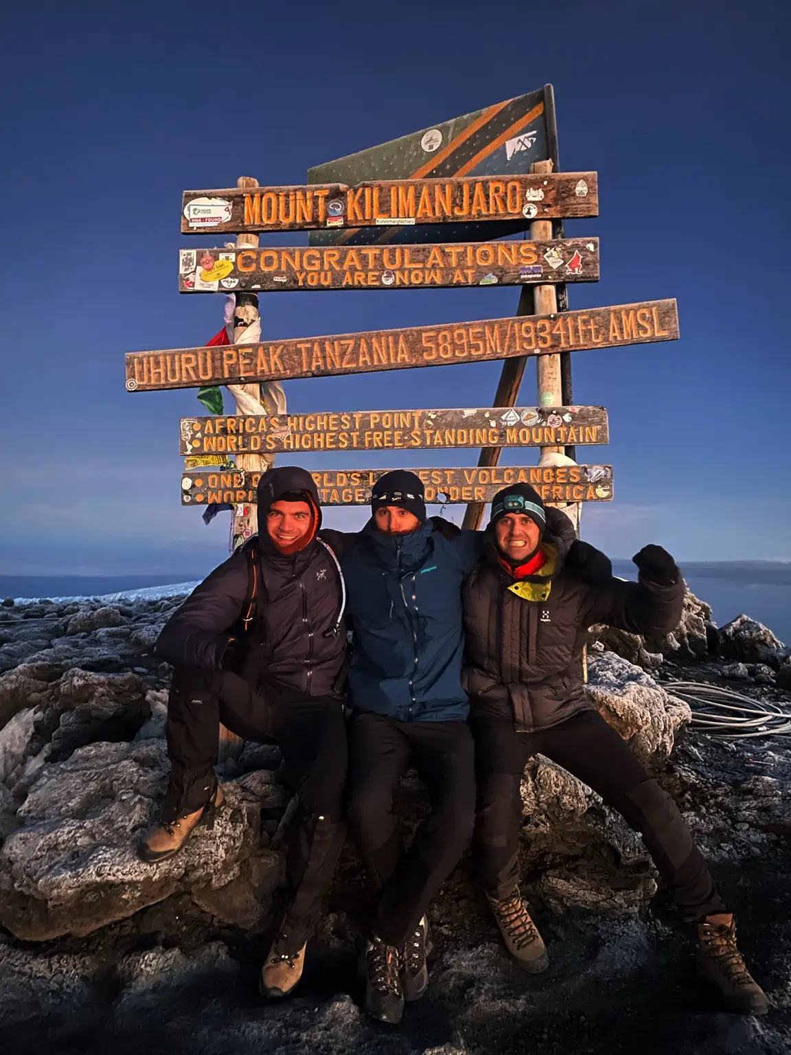 Three expedition members standing at the Uhuru Peak summit sign on Mount Kilimanjaro.